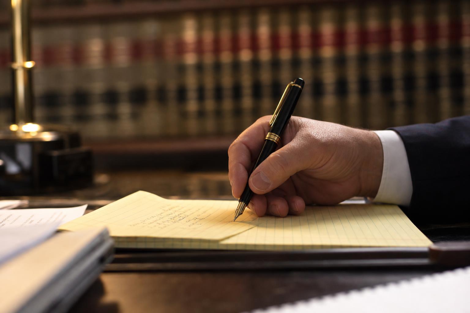 Attorney writing on a yellow legal pad with a fountain pen, law books in background